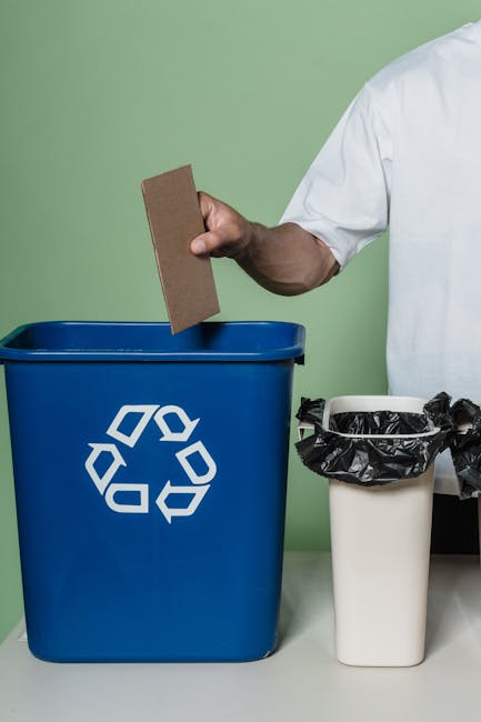A green outdoor trash bin with a black lid and a light blue flap on top, situated on a grassy area in a garden. The bin features a white icon of a person disposal waste into a bin. Surrounding the bin